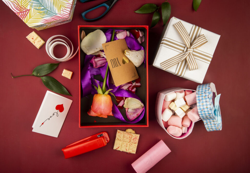 top view of a red and white custom treat boxes with brown paper card and coral color rose flower and petals with purple ribbon and heart shaped box filled with marshmallow on dark red background