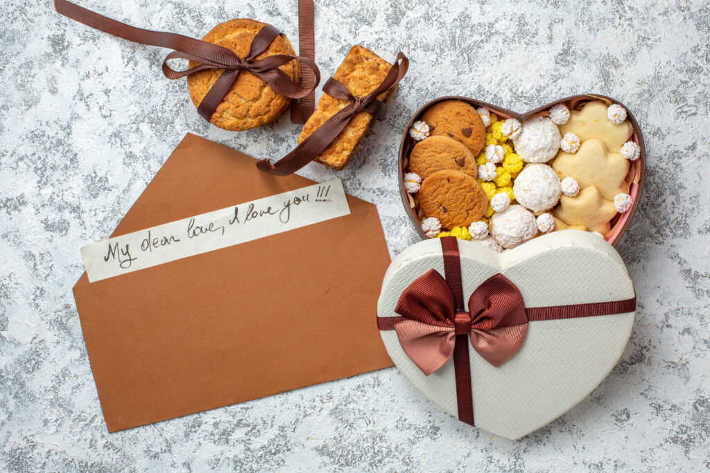 top view of a white color, heart shaped custom treat box with cookies inside and a brown color envelope and two cookies wrapped around a brown color ribbon on a white color design counter top.