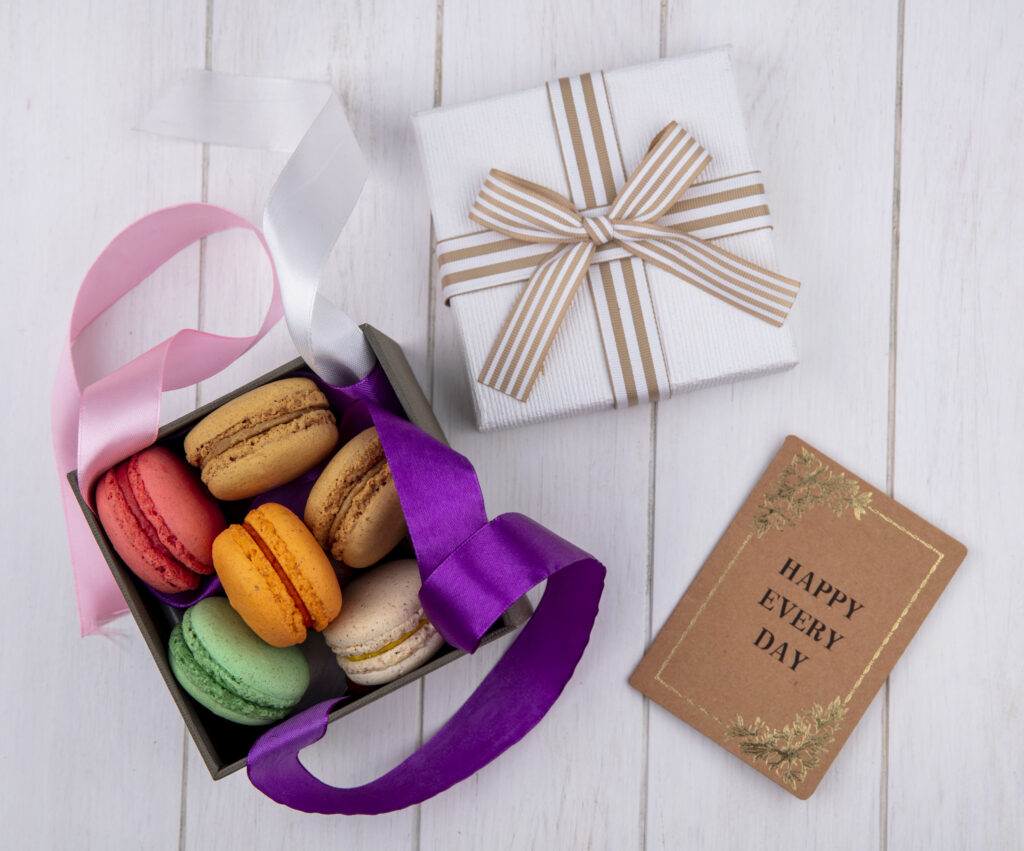 top view colored macarons in a custom macaron box with colored bows and a gift box with a book on a white background
