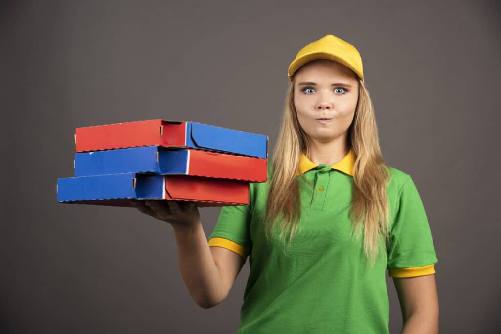 Deliverywoman in uniform holding cardboard compostable packaging of pizza .